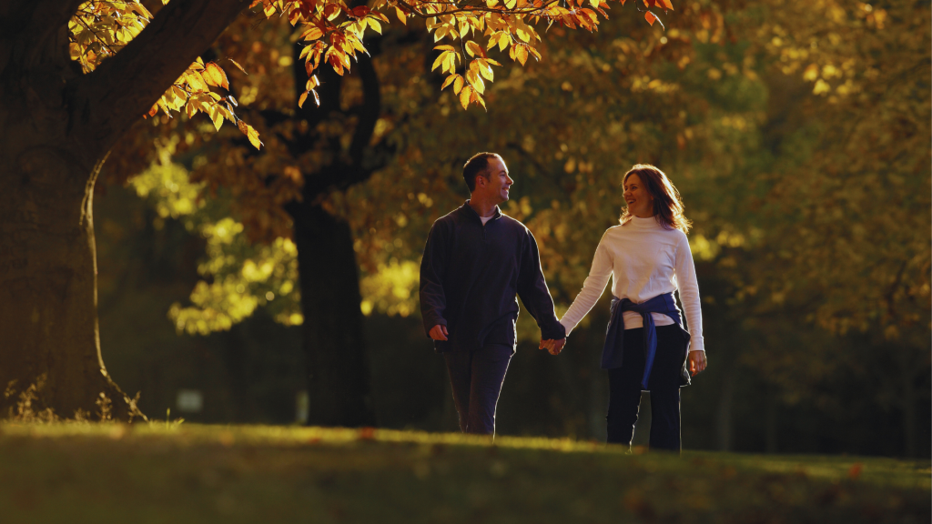 A smiling couple holding hands while walking through a park in autumn.